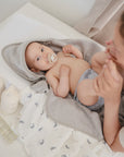 A baby lies on a changing table wrapped in mushie’s gray organic cotton hooded towel, holding a pacifier. An adult gently touches the baby’s feet, and mushie Organic Cotton Muslin Swaddle Blanket is visible nearby.