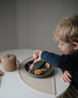A young child sits at a round table, using a pink fork to pick up orange slices from a divided plate on the mushie Silicone Placemat. Nearby, a lidded cup and spoon rest in the softly lit, minimal setting.