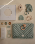 A flat lay of pastel green and beige baby items, featuring a mushie Baby Wrap in its box, a quilted changing mat, pacifiers, a rattle, beanie, stacking toy, teether, and a bottle of baby lotion.
