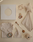A flat lay of beige baby items—onesie, knotted hat, teether rings, pacifier clips, star comforter, mushie Baby Wrap box, rattle, and sippy cup—displayed on a neutral background.