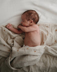A newborn sleeps on a mushie Organic Cotton Muslin Swaddle Blanket, featuring light colors and small rainbow patterns, lying on its side with one arm bent and the other tucked under its face.