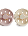 The FRIGG Moon Natural Rubber Pacifier 2-Pack by FRIGG features pale pink and cream pacifiers with moon and star cutouts on the shields and natural rubber latex nipples, shown side by side on a white background.