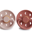 The FRIGG Moon Natural Rubber Pacifier 2-Pack includes two pacifiers—one light pink, one muted red—each with round shields featuring three moon-shaped cutouts and the brand name FRIGG at the bottom.