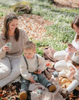 Two women and two toddlers enjoy a picnic outdoors, surrounded by leaves and white flowers. The children play with toys and snack from the spill-proof mushie Snack Cup while the adults relax with drinks nearby.