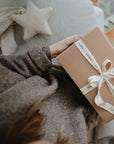 A person in a cozy brown sweater holds a gift wrapped in brown paper with a white ribbon. In the background, the mushie Star Lovey Blanket and a patterned rug are visible.