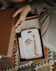 Lifestyle image looking into a Mushie gift box in a person's lap as they lift the lid off the box, revealing a Shifting Sand Star Training Toothbrush and Baby Shampoo in full packaging.