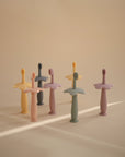 Six mushie Flower Training Toothbrushes with pastel colors and flower-shaped bases stand upright on a beige surface in soft light.