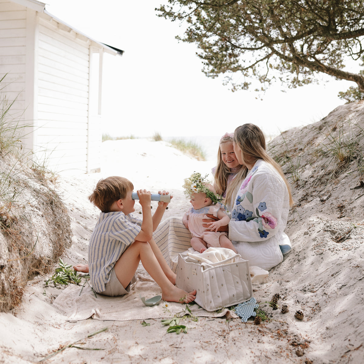 A woman and three kids picnic with a mushie Diaper Caddy on sand near a white cabin and trees.