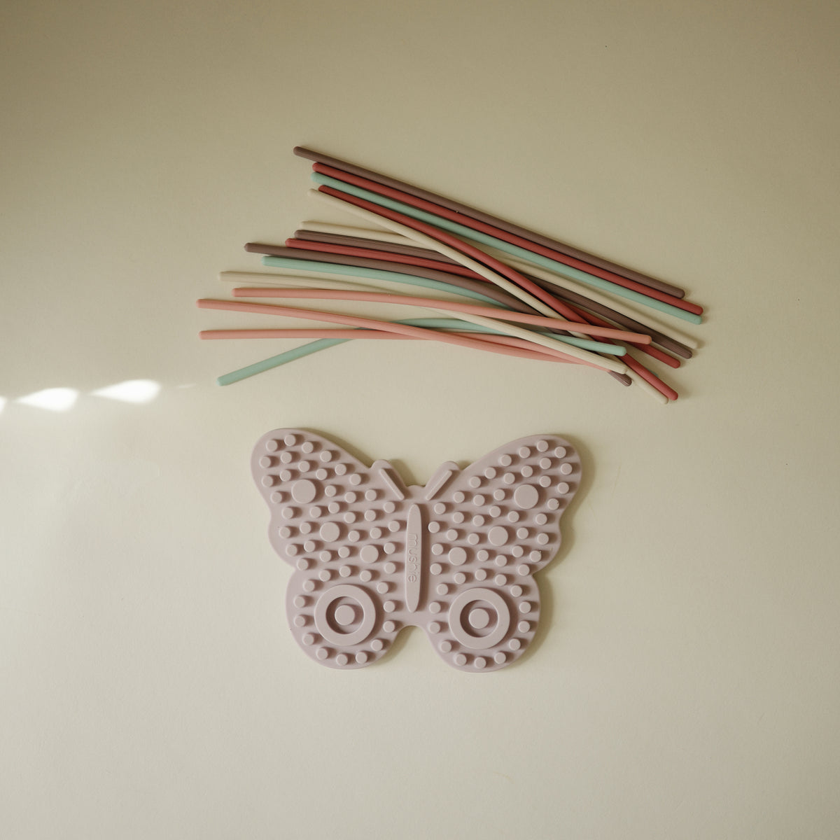 Mushie Silicone Sensory Toy, pink butterfly, under pastel silicone strings on beige surface.