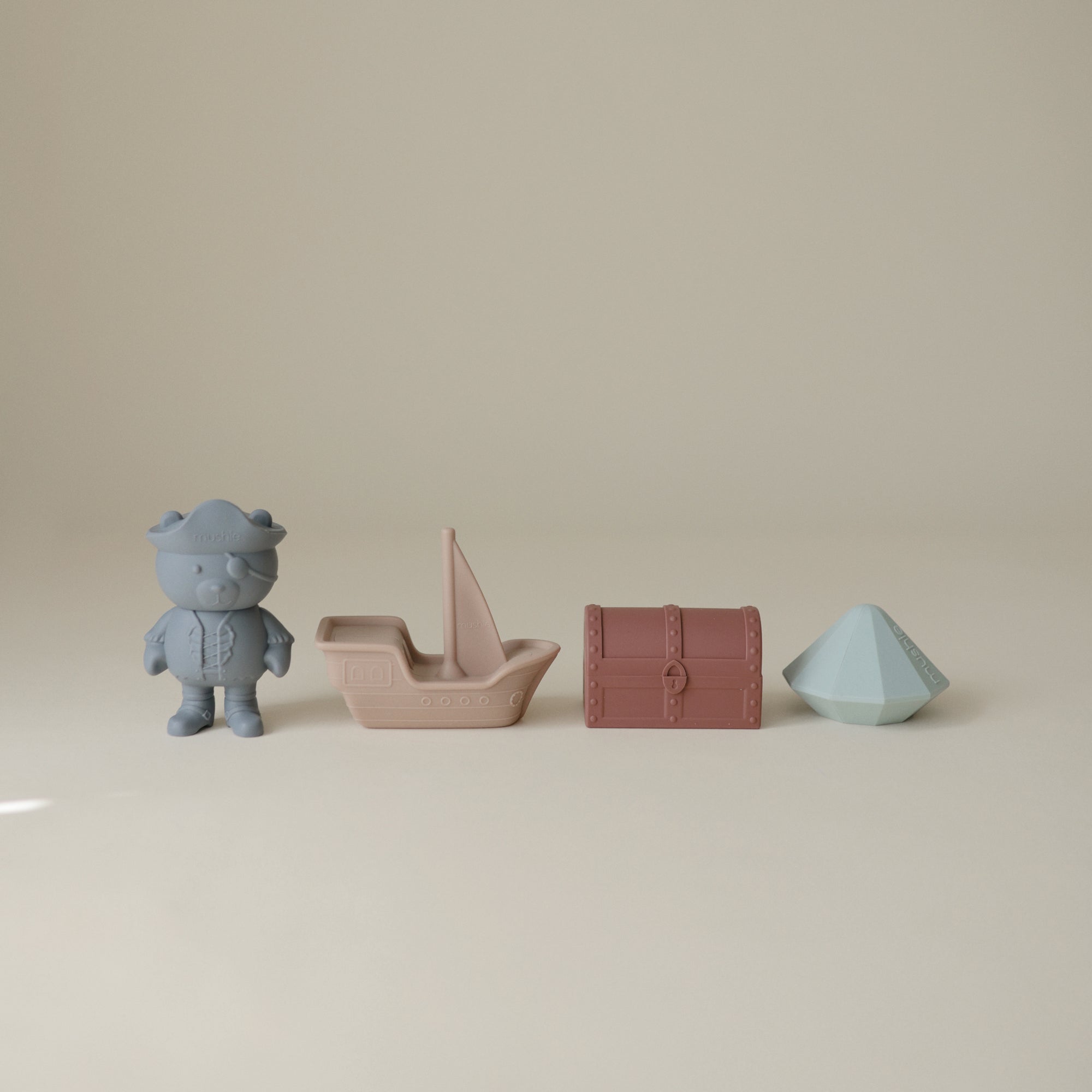 Lifestyle images of a set of Mushie's silicone Pirate Bath Play Set, including a bear, boat, a diamond, and treasure chest on a beige background