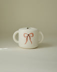 Mushie Snack Cup: spill-proof, white, two handles, pink bow design, plain background.
