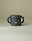 Mushie Snack Cup: Spill-proof silicone, two handles, dark design with planet print for toddlers.
