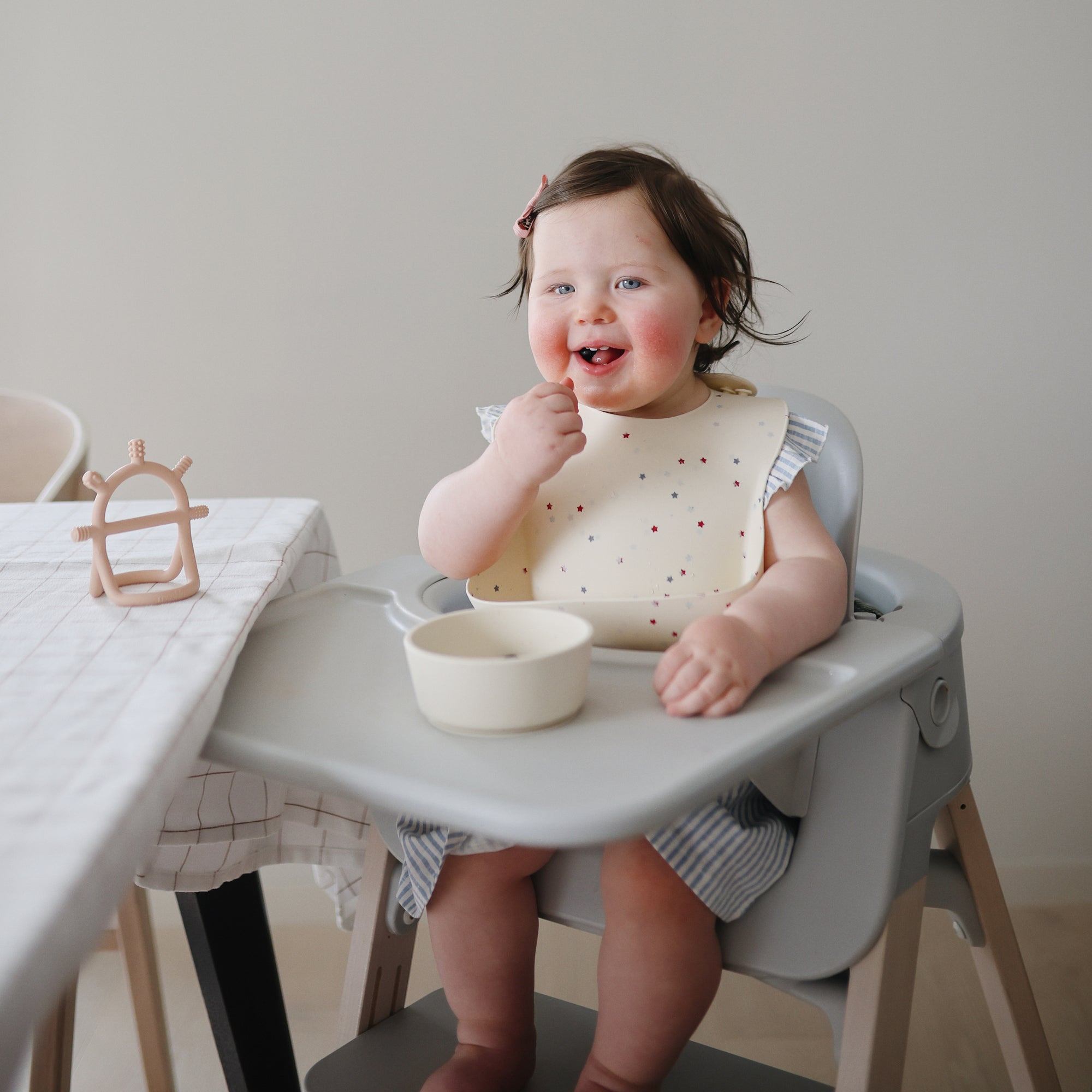 Smiling toddler in high chair wears mushie Silicone Baby Bib with adjustable neck strap.