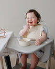 Smiling toddler in high chair wears mushie Silicone Baby Bib with adjustable neck strap.