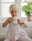 Happy child drinks from Mushie Silicone Training Cup + Straw by a window with plants.