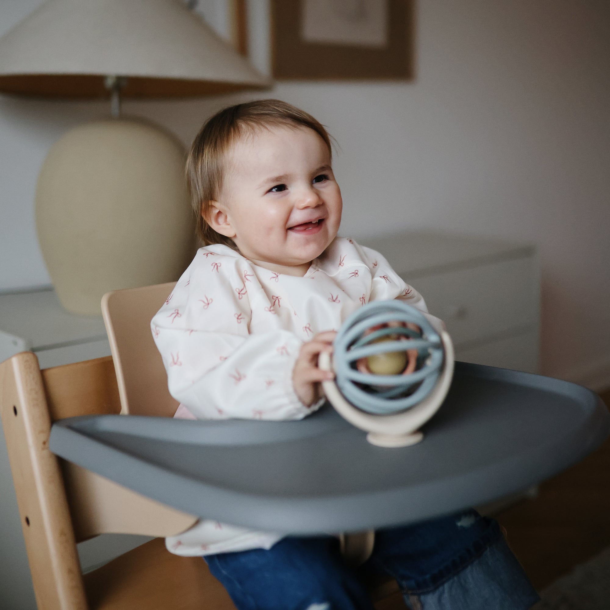 Child sitting in a high chair holding a suction spinner ball toy, smiling.