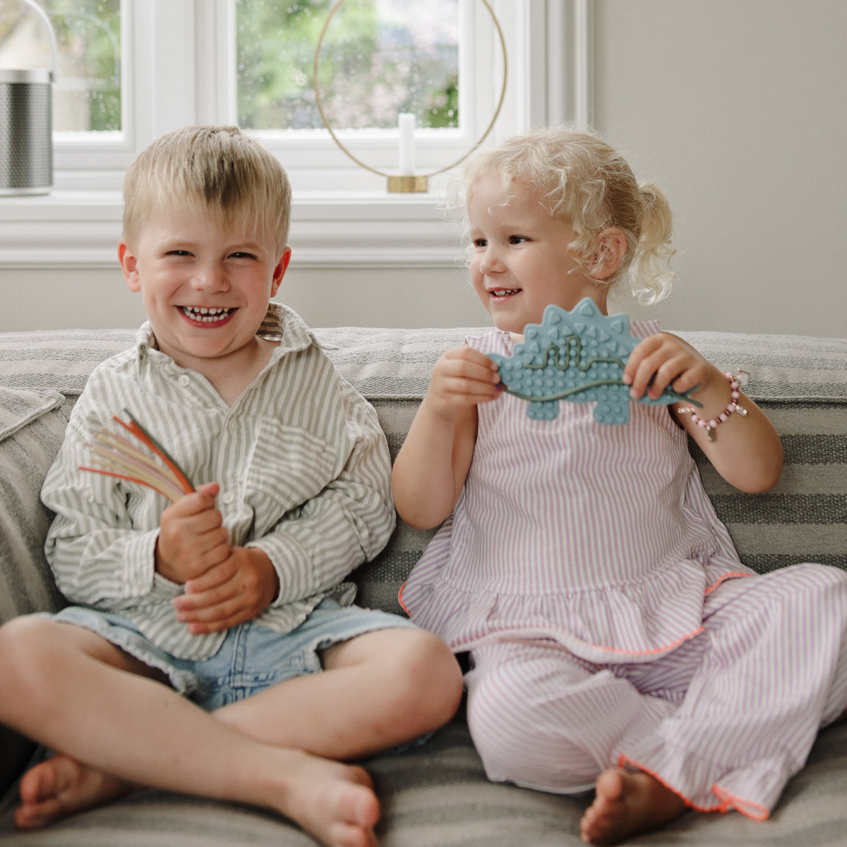 Two smiling kids on a couch: one holds colored pencils, the other a mushie Silicone Sensory Toy.