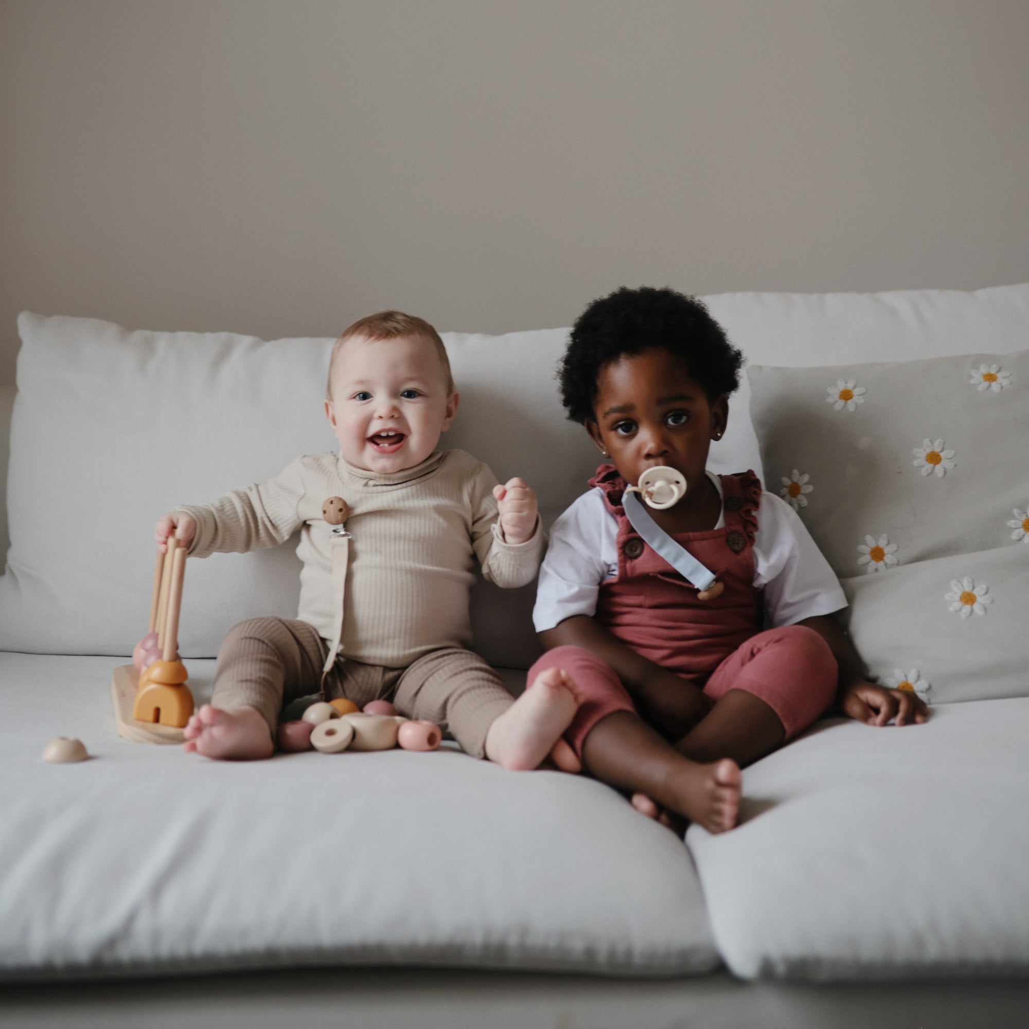 Two toddlers on a couch—one smiles with a toy, the other holds a mushie Linen Pacifier Clip 2-Pack.