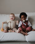 Two toddlers on a couch—one smiles with a toy, the other holds a mushie Linen Pacifier Clip 2-Pack.