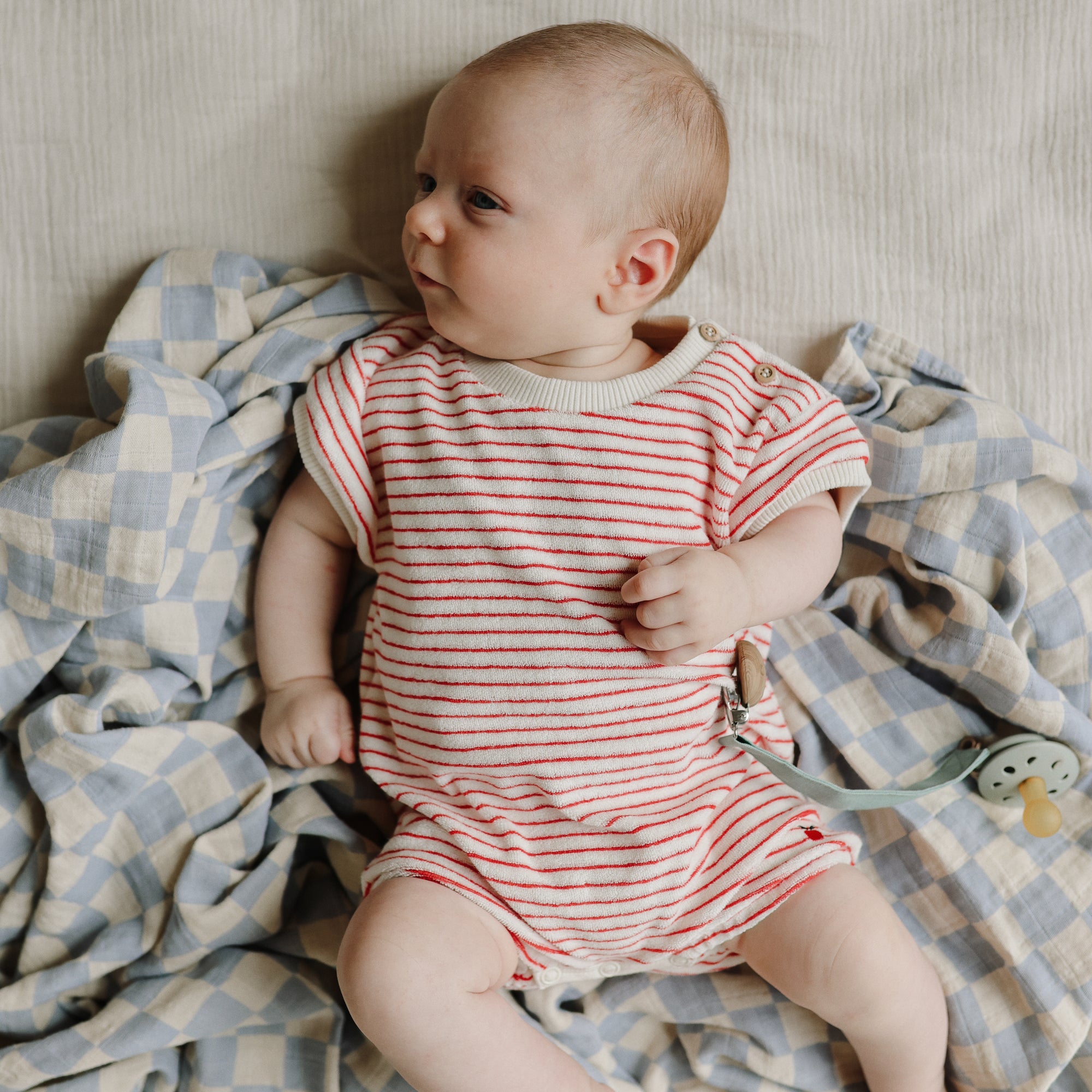 Baby in stripes with mushie Linen Pacifier Clip 2-Pack, silicone clasp, on the blanket.