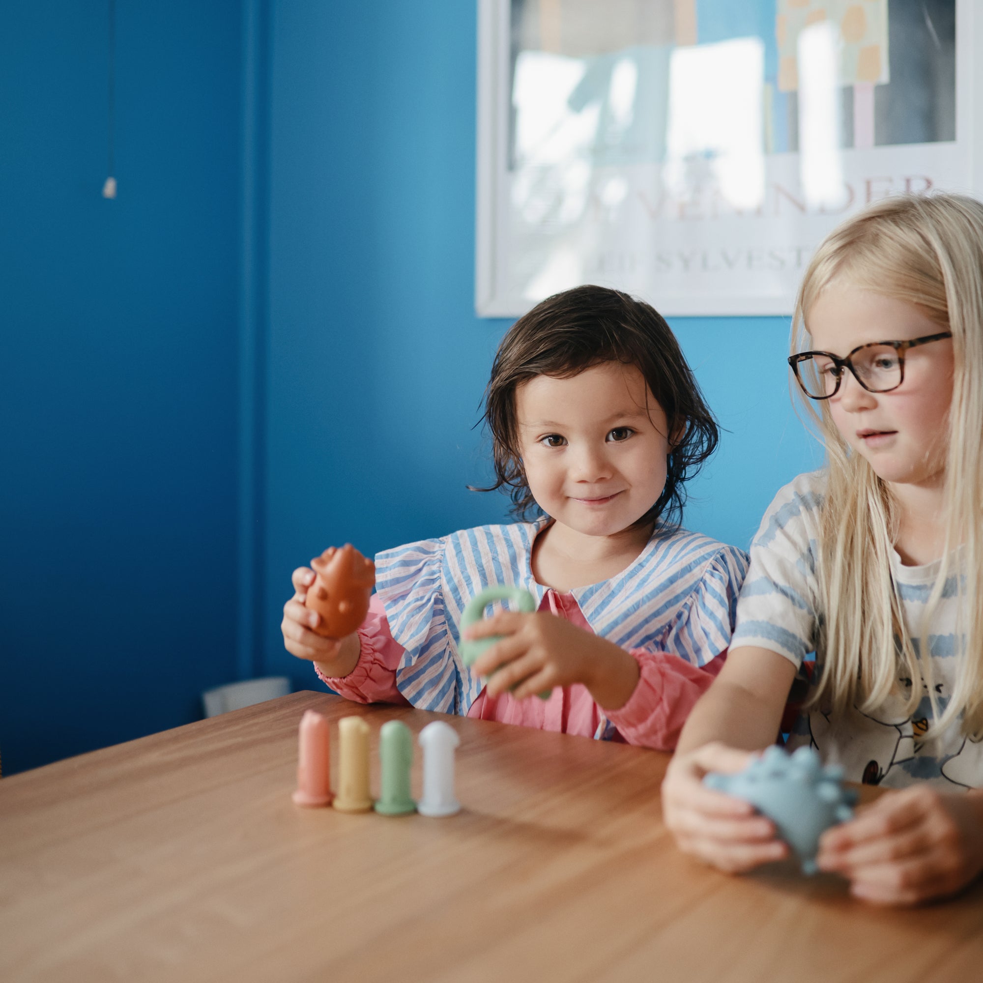 Two kids play with the mushie Mold Free Bath Play Set at a wooden table in a blue room.