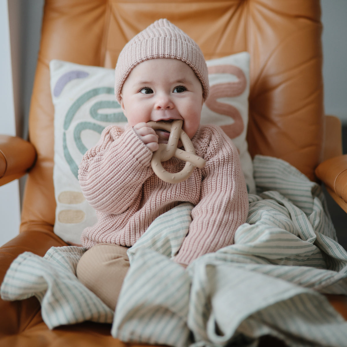 Baby in pink sweater holds mushie Teething Gift Set, smiling while sitting on a chair.
