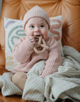Baby in pink sweater holds mushie Teething Gift Set, smiling while sitting on a chair.