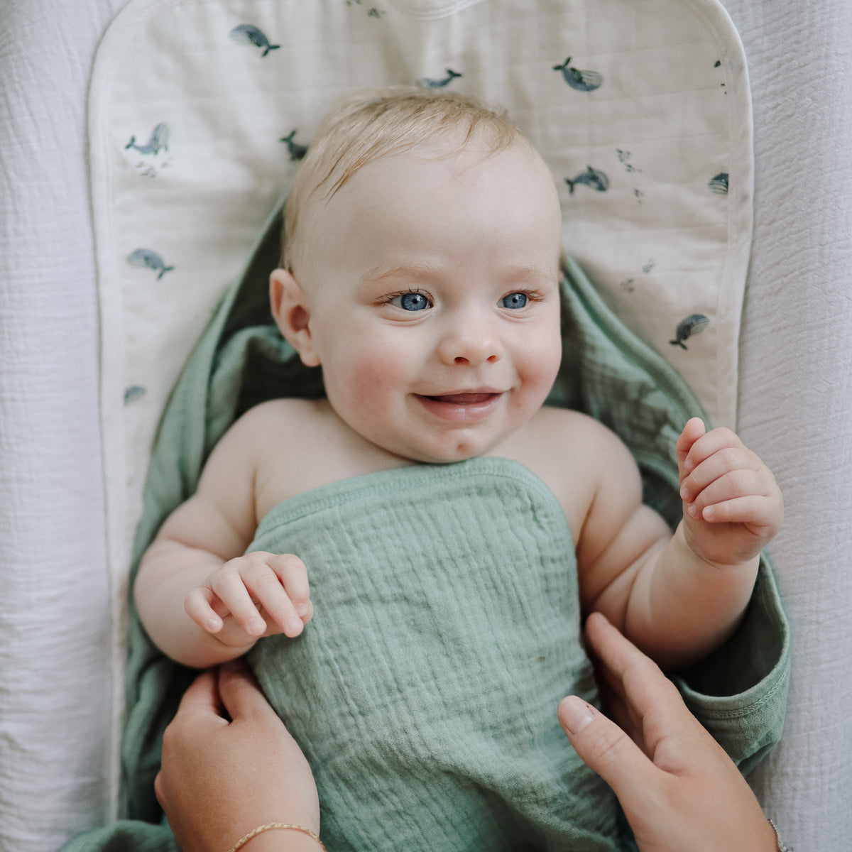 A smiling baby wrapped in sage green muslin on a Mushie Changing Pad Liner.