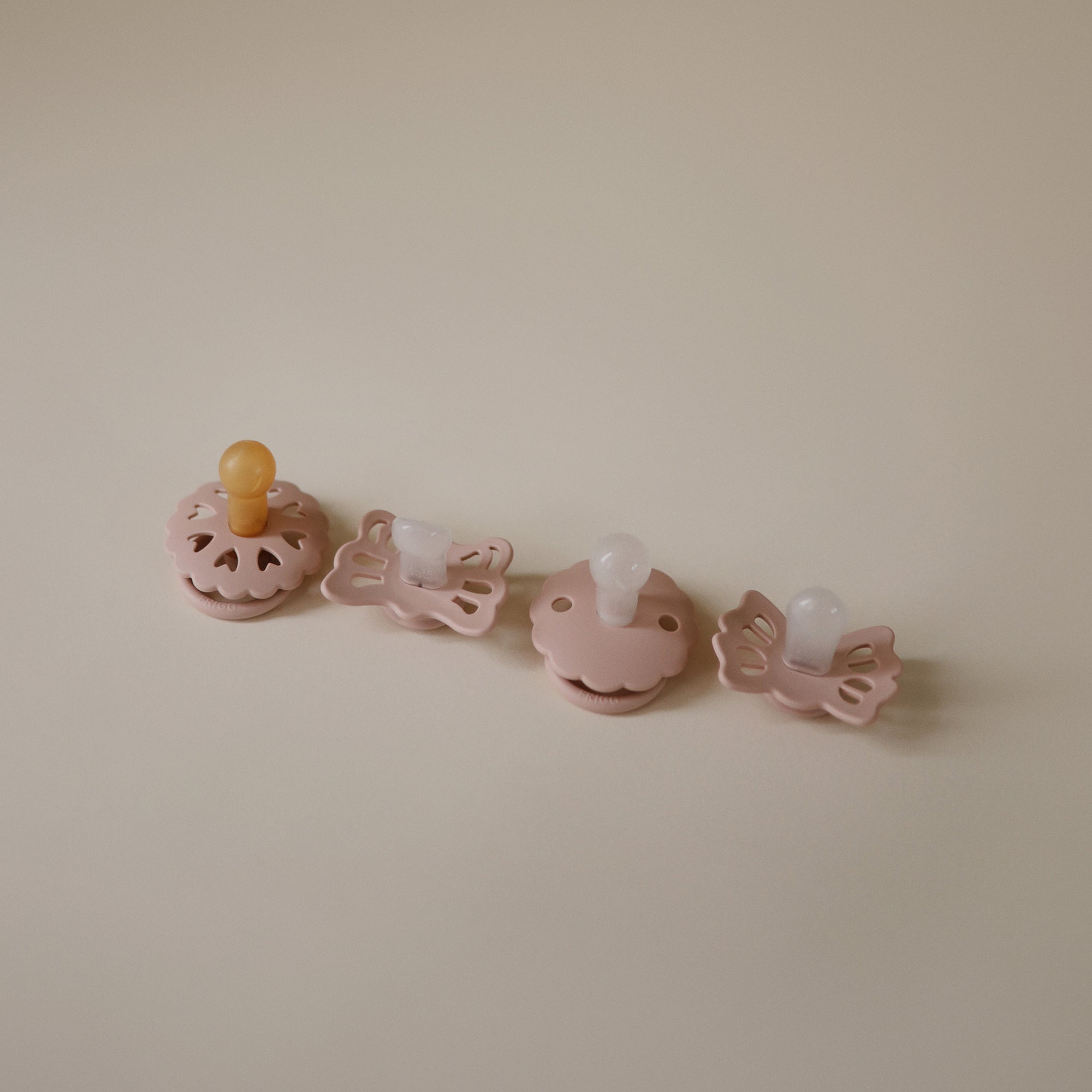 Set of four Blush baby pacifiers on a beige background. 