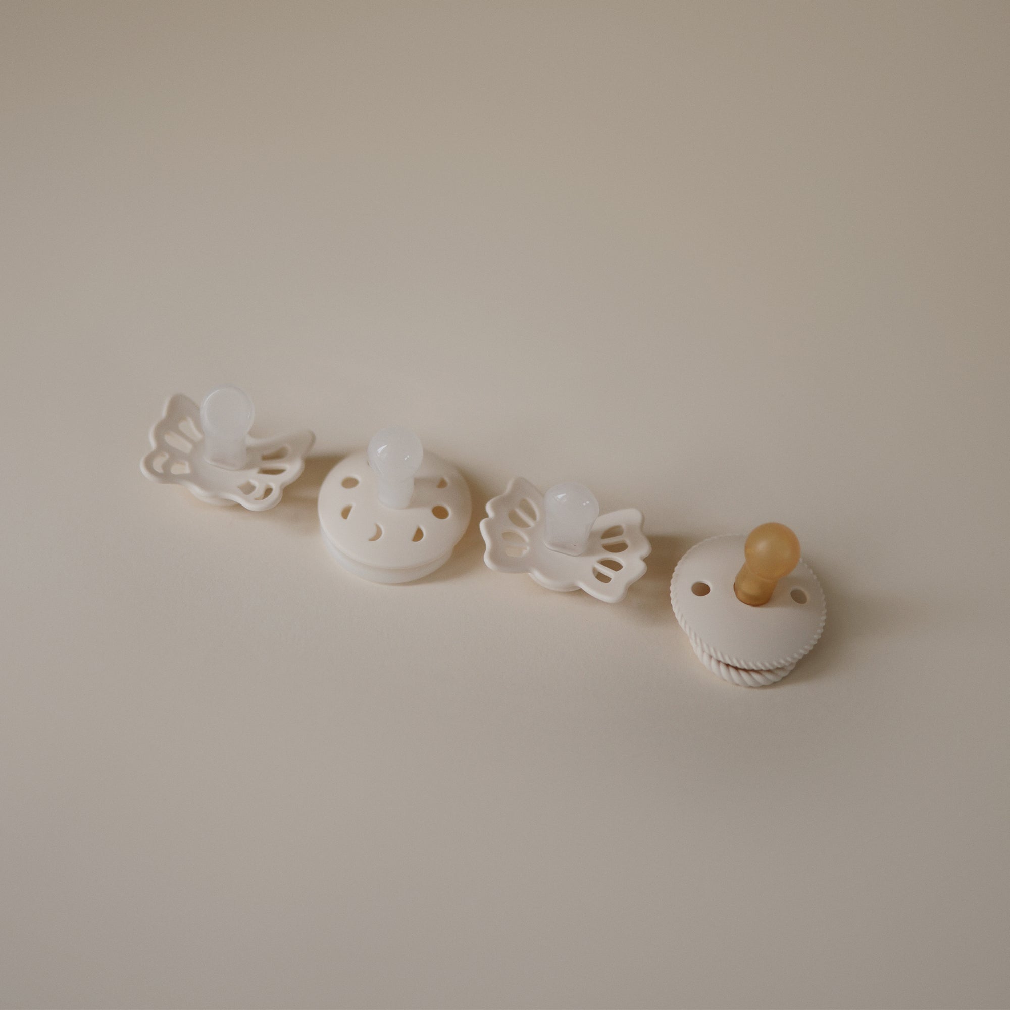 The FRIGG Baby's First Pacifier Moonlight Sailing (Cream) 4-Pack, by FRIGG, features four cream pacifiers in various shapes with silicone nipples, neatly arranged on a light beige surface.