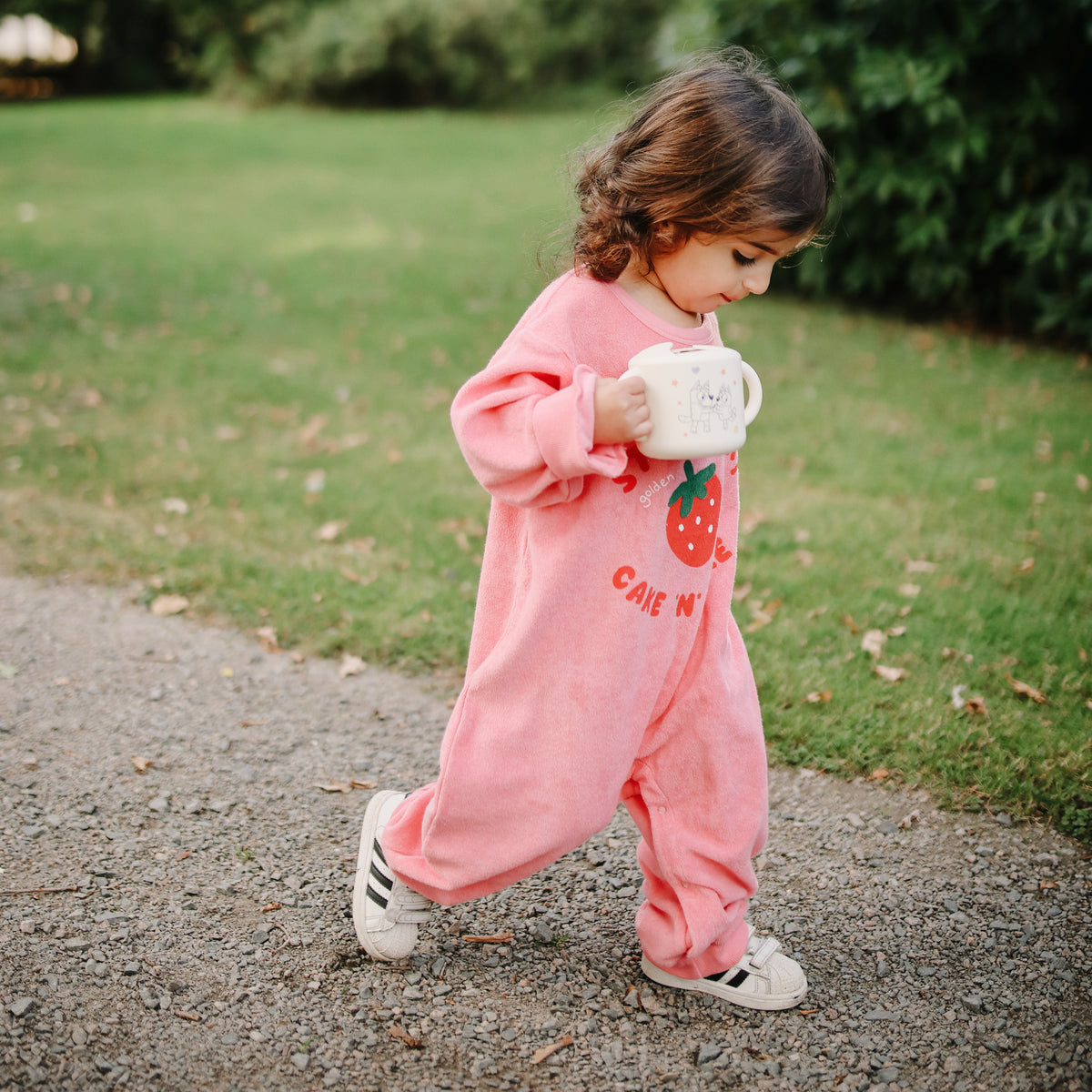 Toddler in pink onesie walks outside holding a mushie Snack Cup in both hands.