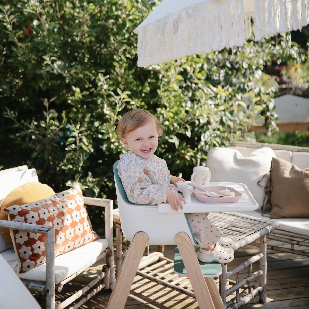 Smiling toddler outdoors, sipping from Mushie Silicone Training Cup + Straw in high chair.