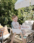 Smiling toddler outdoors, sipping from Mushie Silicone Training Cup + Straw in high chair.