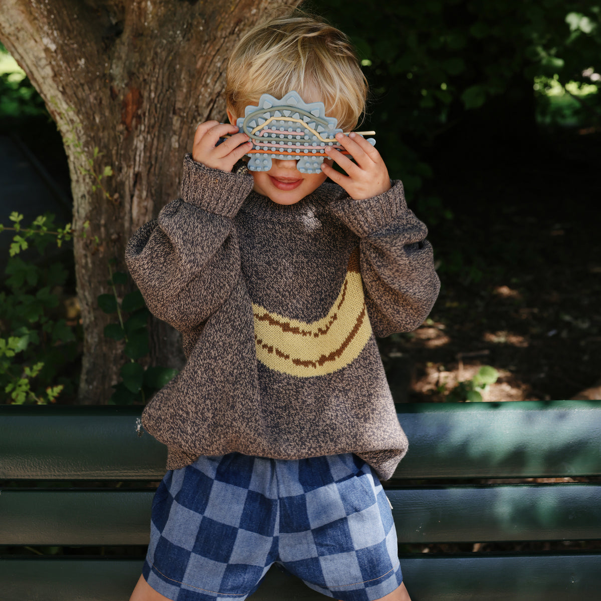 Child outside in a sweater and shorts holds mushie Silicone Sensory Toy over their face.