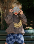 Child outside in a sweater and shorts holds mushie Silicone Sensory Toy over their face.