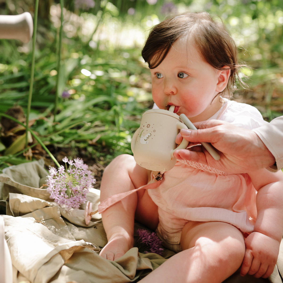 A baby in pink uses the Mushie Silicone Training Cup + Straw on a blanket near purple flowers.