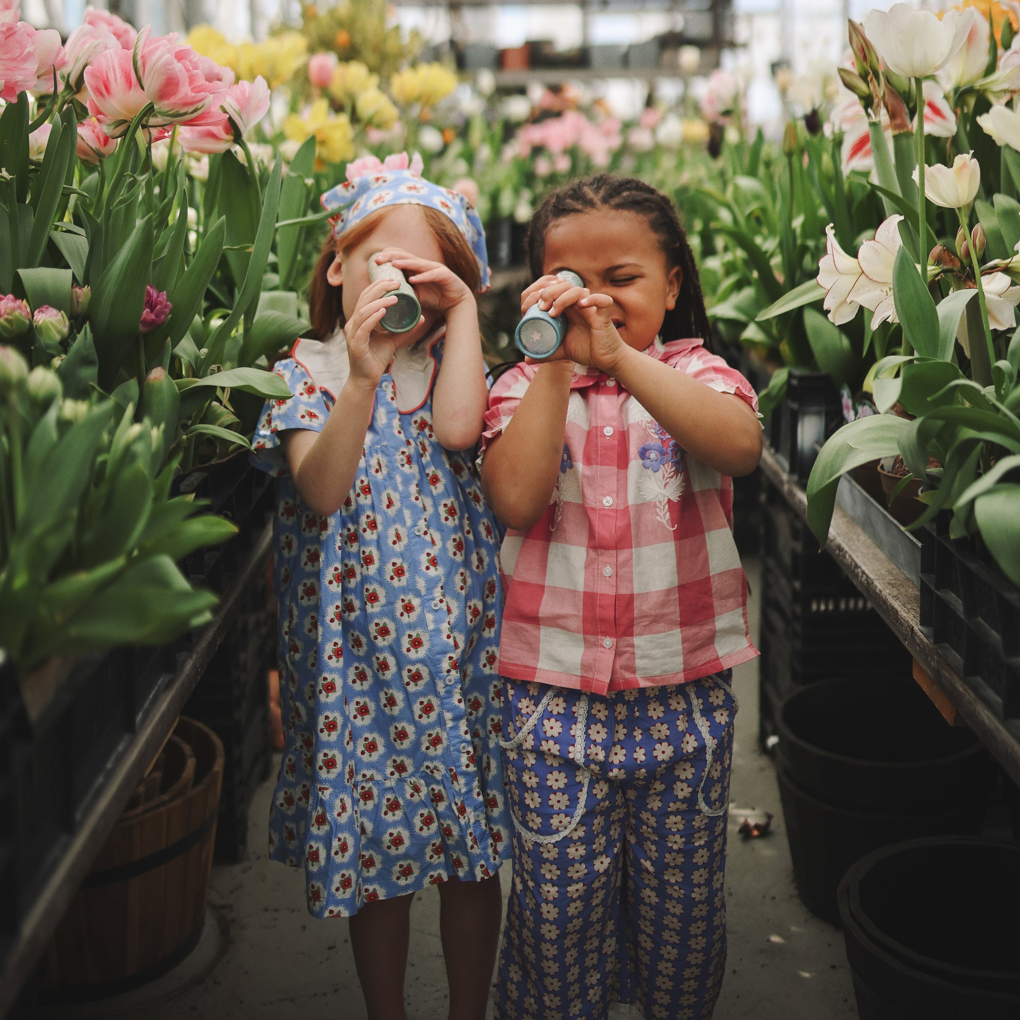 Two kids play with mushie Kaleidoscopes among flowers in a greenhouse.