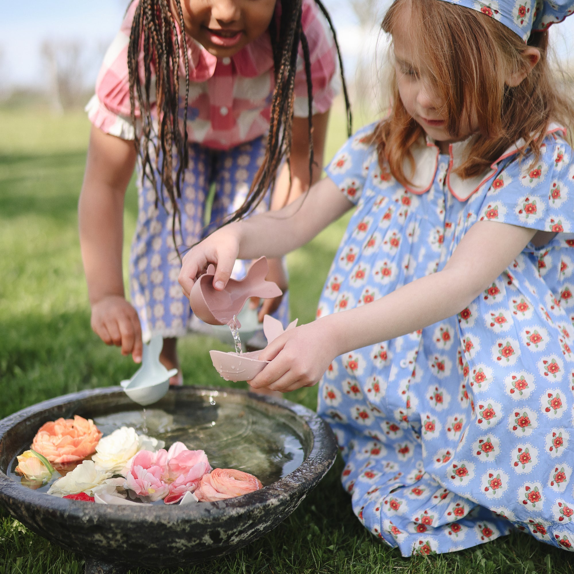Two young girls pour water into a birdbath with Mushie Whale Bath Stackers on the lawn.