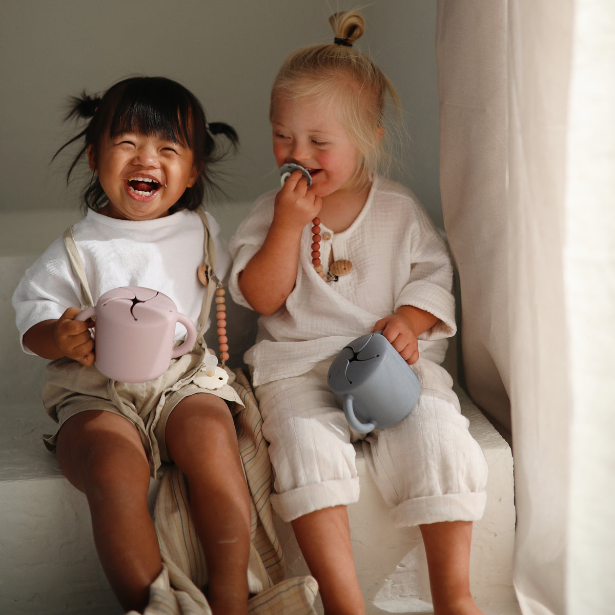 Two toddlers laugh with pastel mushie Snack Cups, sitting on white steps by a window.
