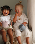 Two toddlers laugh with pastel mushie Snack Cups, sitting on white steps by a window.