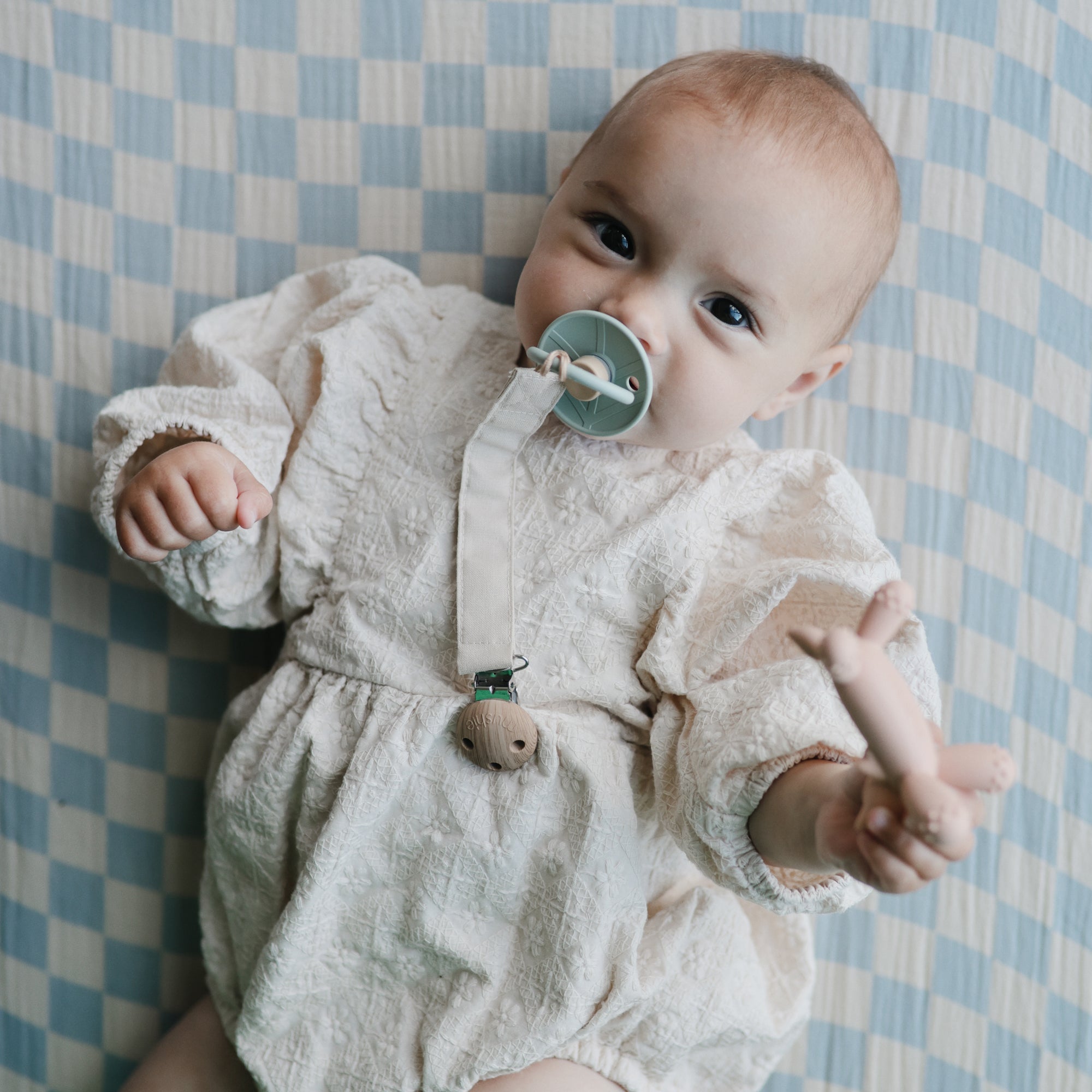 Baby with mushie Linen Pacifier Clip 2-Pack on checkered blanket, wearing romper.