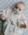 Baby with mushie Linen Pacifier Clip 2-Pack on checkered blanket, wearing romper.