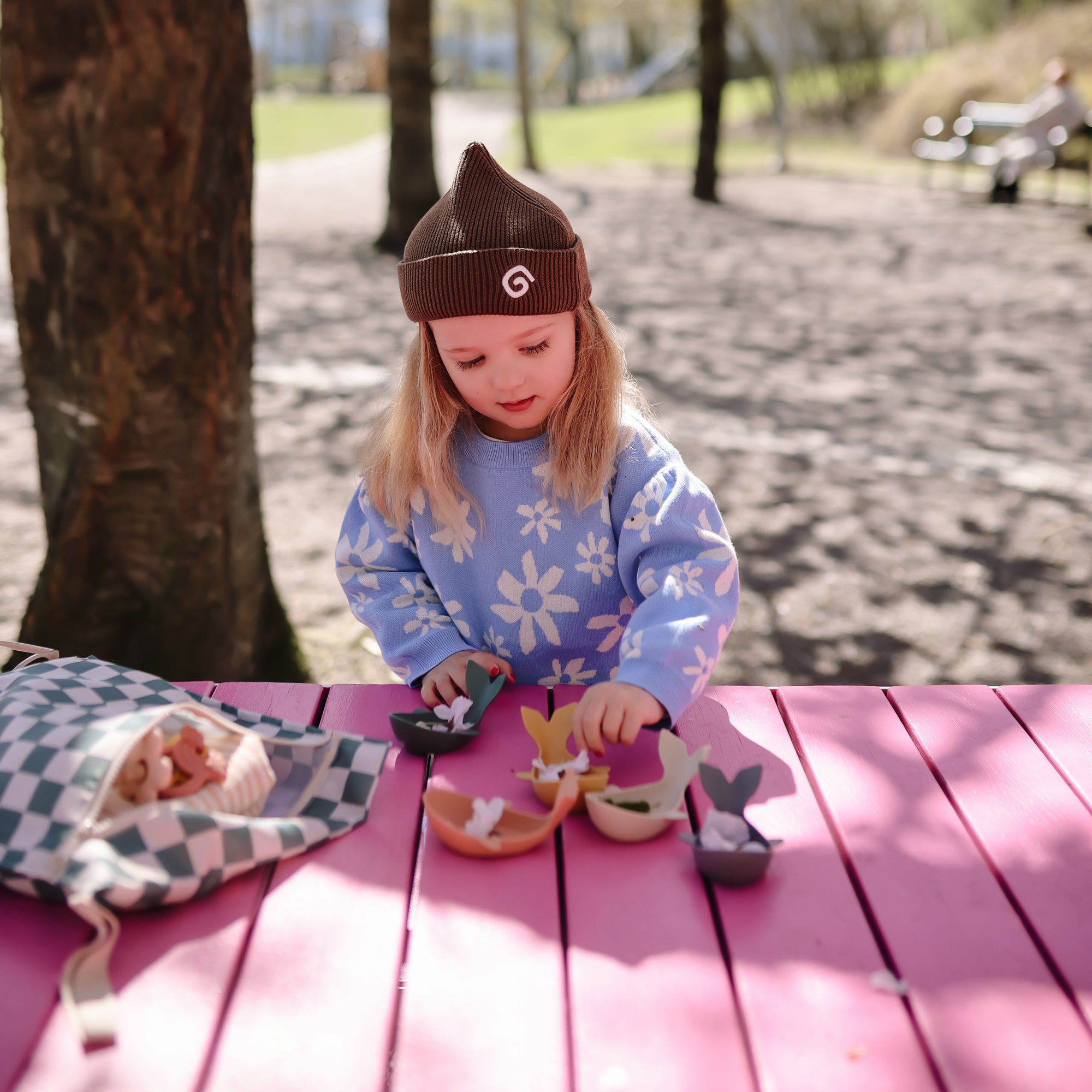 A young girl plays with Mushie Whale Bath Stackers on a pink table in a sunny park.