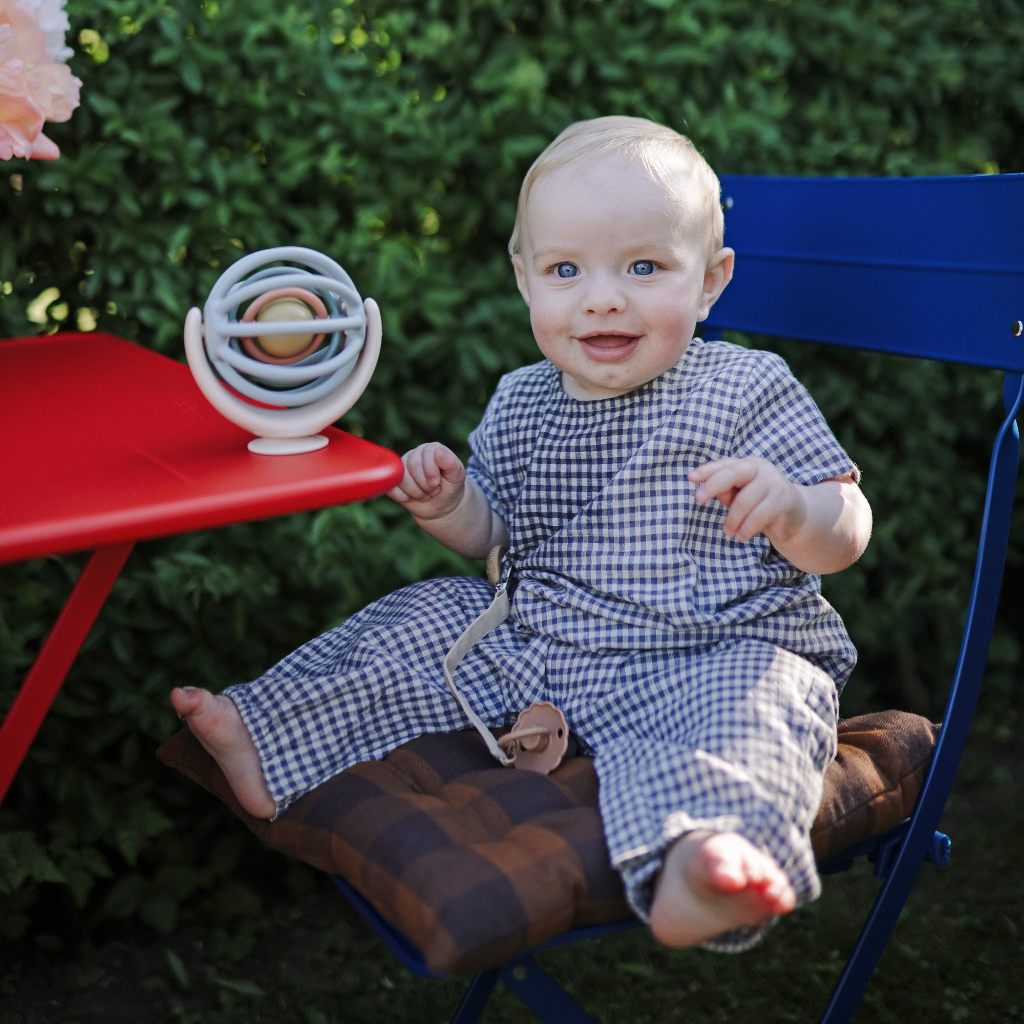 Baby in gingham with mushie Linen Pacifier Clip 2-Pack sits on blue chair by globe outdoors.