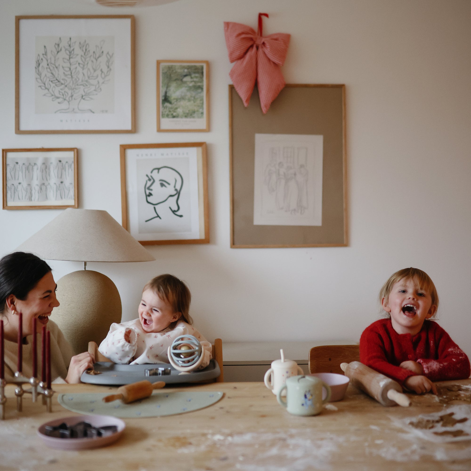 A woman and two young children smile and laugh at a table covered with baking supplies, brightened by Bluey x Mushie Silicone Placemats from mushie. Behind them, the wall features framed art and a big red bow.