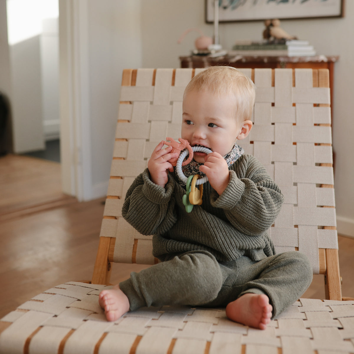Toddler in green outfit on chair chews a mushie Teething Gift Set sensory teether.