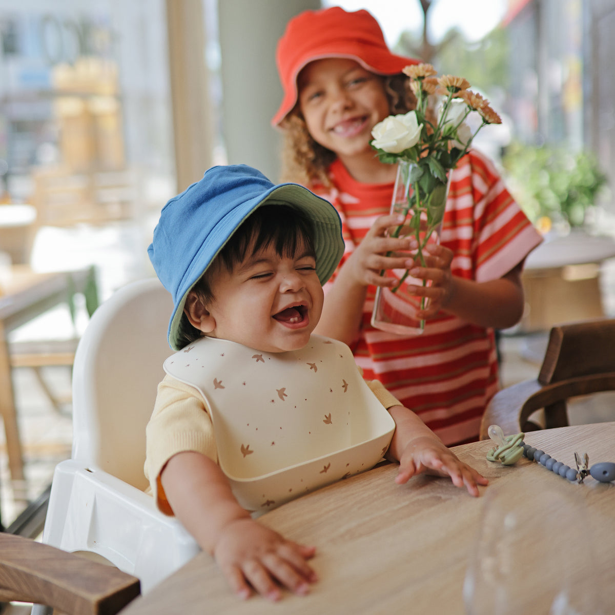 Smiling toddler in blue hat wears mushie Silicone Baby Bib; older child holds flowers.