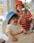 Smiling toddler in blue hat wears mushie Silicone Baby Bib; older child holds flowers.