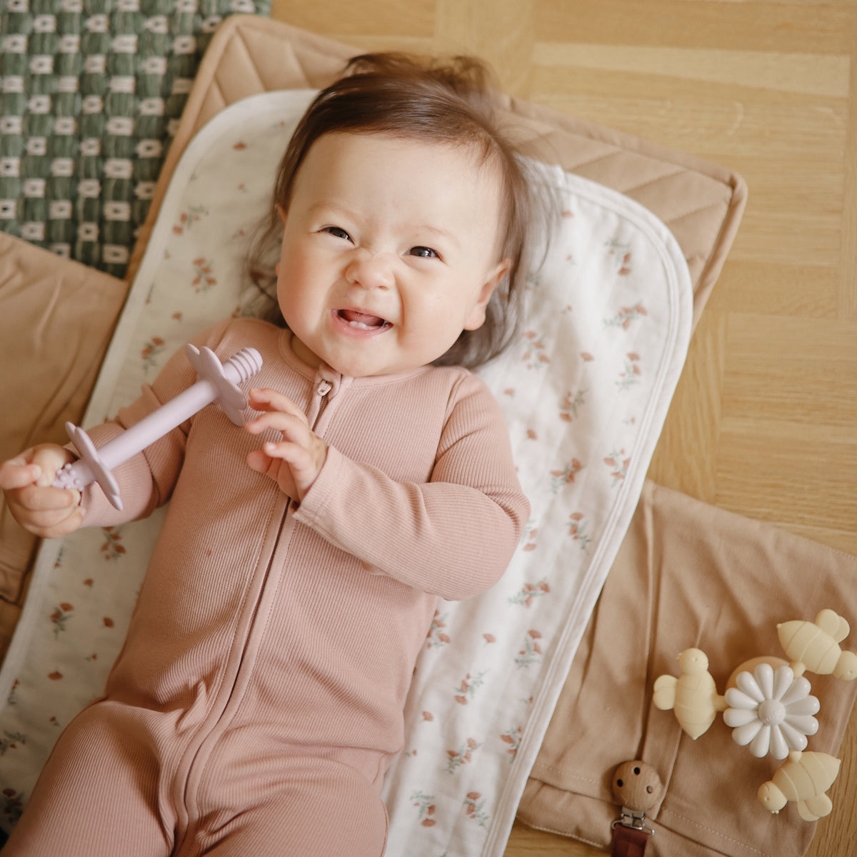 A smiling baby holding a teether lies on a floral Mushie Changing Pad Liner.