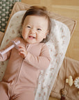 A smiling baby holding a teether lies on a floral Mushie Changing Pad Liner.